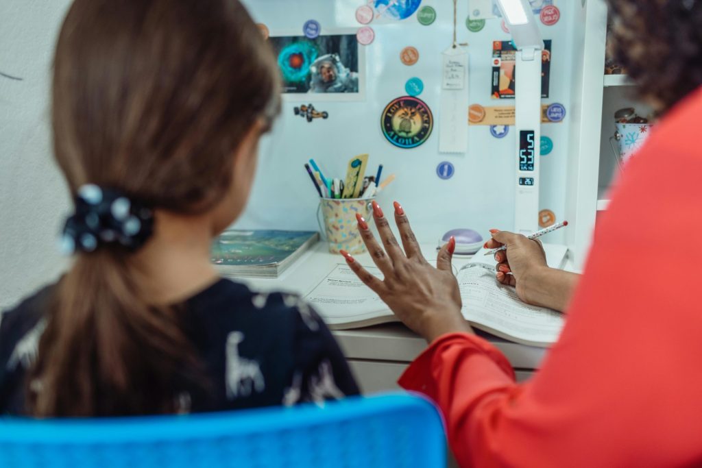 Crop anonyme ethnische weibliche Tutorin im Gespräch mit Schulmädchen am Tisch mit Übungsbuch im Haus